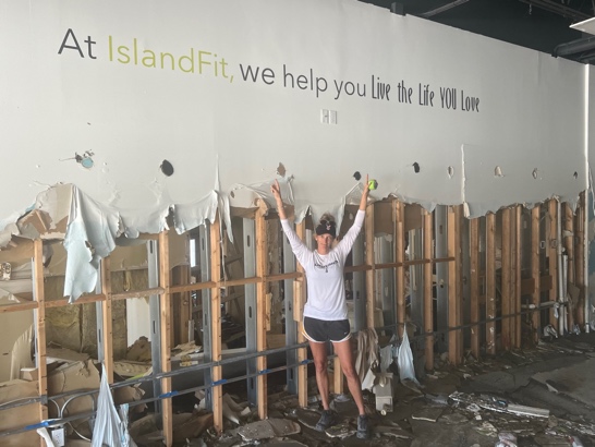 An IslandFit team member celebrating in front of the storm-damaged wall that reads At IslandFit, we help you live the life you love
